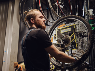 Fototapeta na wymiar Handsome redhead male in a jeans coverall, working with a bicycle wheel in a repair shop. A worker removes the bicycle tire in a workshop.