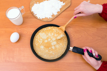 Wonam holding a frying pan with traditional russian pancake. A glass of milk, an egg and a cutting board with flour are on the wooden table