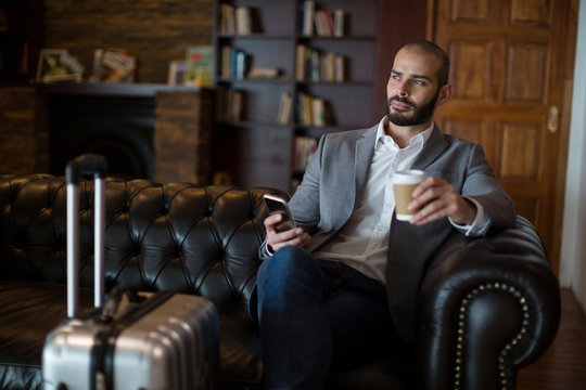 Thoughtful businessman holding mobile phone and coffee cup in waiting area