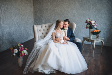 Beautiful newlyweds sitting on sofa in gray studio and hugging. A modern bride and groom in a lace...