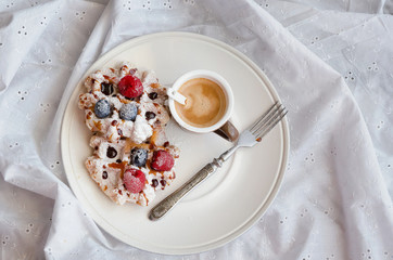 Cup of black coffee and homemade belgian waffles with berries and icing sugar on plate on bed. Good Morning dessert background. Top view.