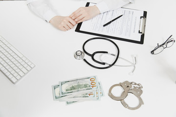 Close up hands female doctor with medical documents at desk in hospital office. Woman in medical gown, handcuffs, bundle of dollars, cash money on table in consulting room. Medicine, law concept.