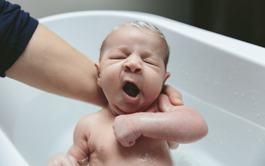 Newborn girl yawning in the bathtub held by her mother