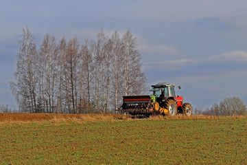 A farmer on a red tractor with a seeder sows grain in plowed land in a private field in the village area. Mechanization of spring field work. Farmer's everyday life. Processing of land. The agrarian.