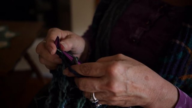 Close-up of retired woman knitting some purple yarn while sitting with her husband enjoying their hobbies.