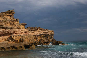 An opening in the storm clouds allows a moment for the sunlight to shine on the rocks of London Bridge in Portsea, Victoria, Australia