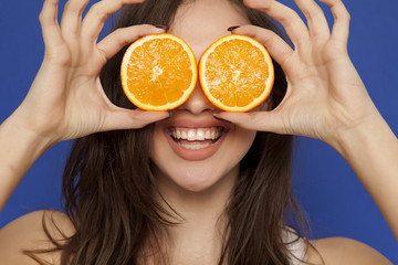 Happy young woman posing with slices of oranges on her face on blue background