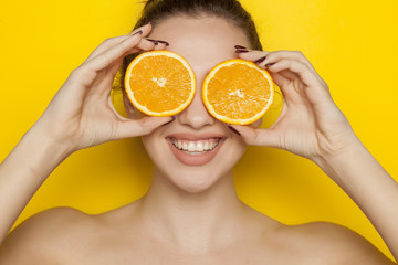 Happy young woman posing with slices of oranges on her face on yellow background