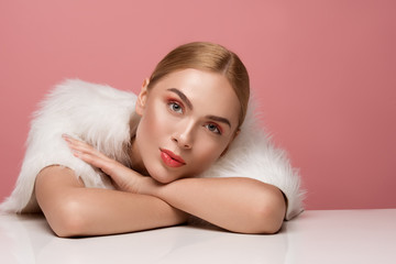 Portrait of appealing woman in fur coat sitting at the table. Her head reposing on arms. Copy space in right side. Isolated on rose background