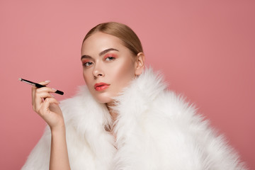 Portrait of gorgeous serene girl wearing fluffy coat. She is holding makeup brush like cigarette. Isolated on rose background