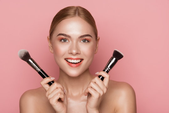 Visage services concept. Portrait of satisfied woman holding one blush brush in each hand and staring at camera with joy. Isolated on rose background