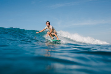 happy young man in wet t-shirt riding waves on surfboard on sunny day