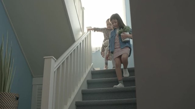 Pan of little Asian girl with potted plant and plush bear toy walking down staircase