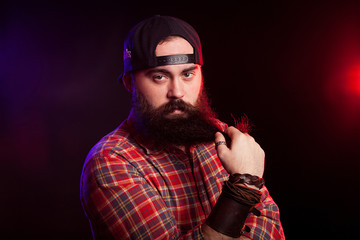 Angry bearded hipster man holding his beard in hands on black background with red and blue lights behind