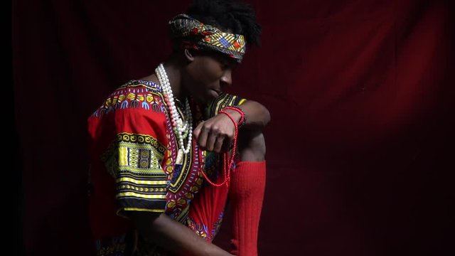 A black man in long socks and a bright dasha of angola sits on a red background, turns on the camera