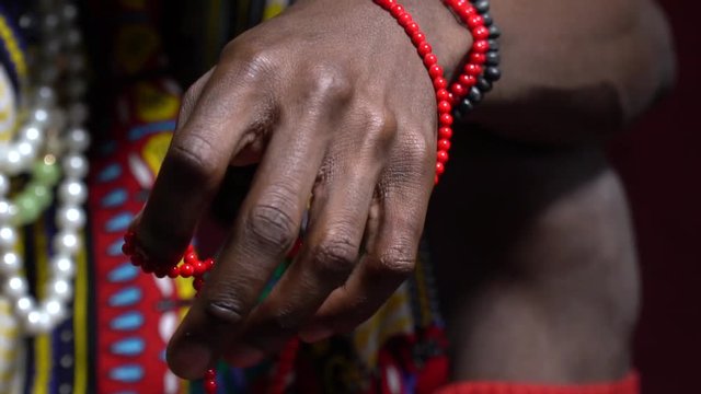 A black male from Angola in national clothes is holding beads while sitting on a red background