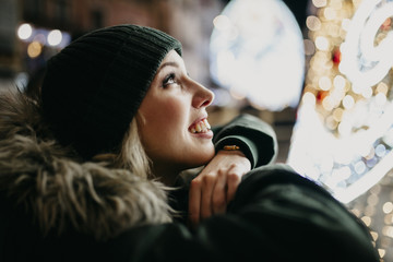 Smiling white woman wearing a green knit hat and coat, looking up and watching city lights during...