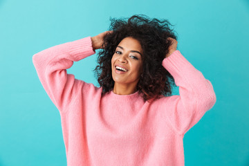 Fototapeta na wymiar Colorful portrait of smiling woman in pink shirt posing on camera and touching her dark afro hairdo, isolated over blue background