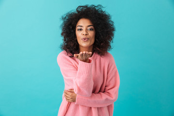 Multicolor image of flirty woman 20s with afro hairstyle smiling and giving air kiss on camera, isolated over blue background - obrazy, fototapety, plakaty