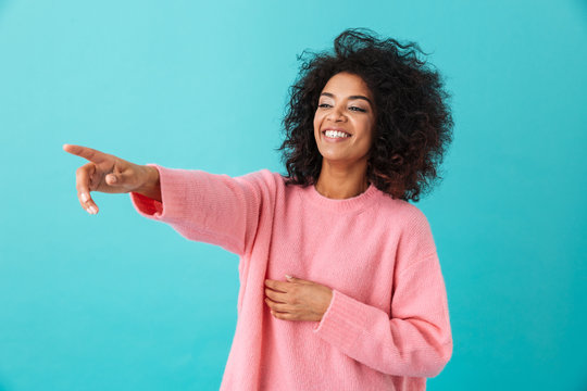 Image of american curly woman in pink shirt looking aside and pointing index finger on copyspace with smile, isolated over blue background