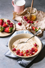 Sweet rice porridge pudding in ceramic plate with berries strawberry and raspberry, walnuts, honey and mug of milk on crumpled paper over grey kitchen table.