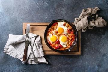Photo sur Plexiglas Oeufs sur le plat Traditional Israeli Cuisine dishes Shakshuka. Fried egg with vegetables tomatoes and paprika in cast-iron pan on wooden board with cloth and cutlery over blue texture background. Top view, space.
