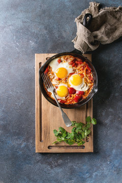 Traditional Israeli Cuisine dishes Shakshuka. Fried egg with vegetables tomatoes and paprika in cast-iron pan on wooden board with cloth and herbs over blue texture background. Top view, space.