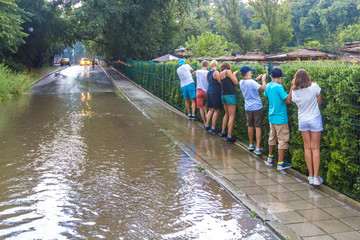 Fototapeta na wymiar Flooding after heavy rain, tourists bypass puddle along edge of sidewalk, following each other in row, holding onto fence, Bulgaria, Golden Sands.