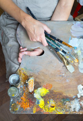 A girl artist in a linen apron holds on her knees a working palette with oil paints and brushes