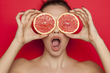 Young surprised woman posing with slices of red grapefruit on her face on red background