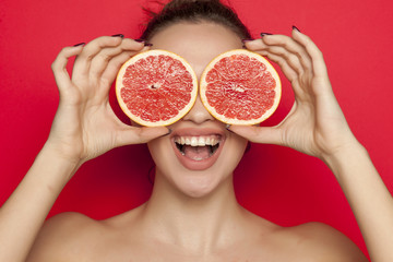 Happy young woman posing with slices of red grapefruit on her face on red background