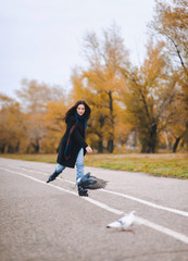 Fototapeta premium A pretty young girl in a coat and blue jeans with a long scarf happily skates in the autumn park with pigeons. Fighting the autumn depression.