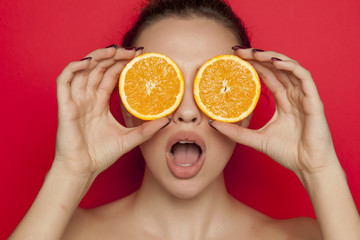 Surprised young woman posing with slices of oranges on her face on red background