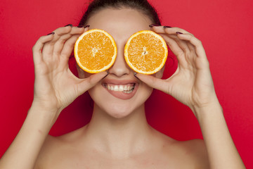 Happy young woman posing with slices of oranges on her face on red background