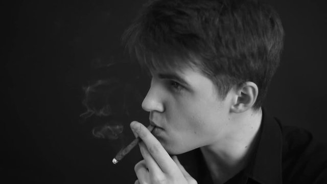 Close-up  caucasian man smoking a cigar. Wearing black  shirt. Studio portrait on  black and white. dark and sullen shot of a young man smoking over a black background