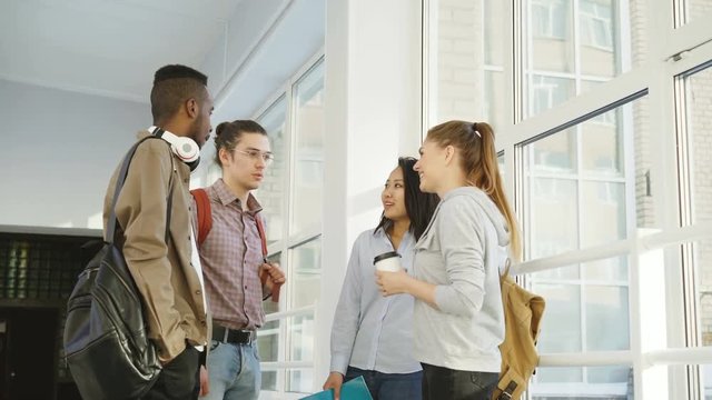 Four multi-ethnic studens are standing in big white spacious hall in college talking to each other in positive way. They are smiling positively and laughing