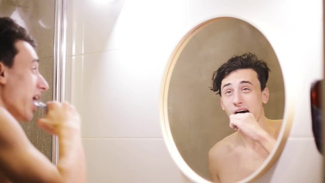 Young man brushing his teeth in bathroom. health care, dental hygiene, people and beauty concept - smiling young man with toothbrush cleaning teeth and looking to mirror at home bathroom