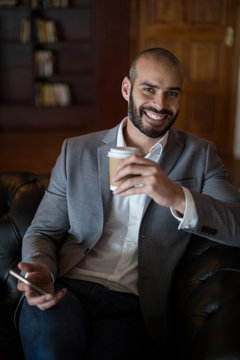 Portrait of smiling businessman holding mobile phone and coffee cup in waiting area