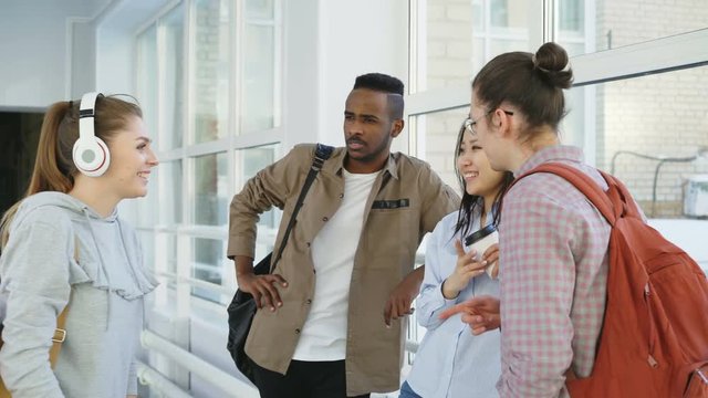 Beautiful attractive female student wearing headphones is going down corridor meeting her groupmates giving five to her friend and moving on. She show middle finger to african american guy