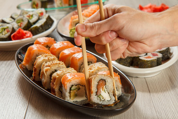 Male hand with two  chopsticks holding Uramaki roll with Conger and different  sushi rolls with seafood on ceramic plates on the background