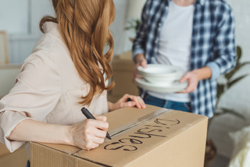 partial view of woman signing cardboard box with husband with dishes near by, moving home concept