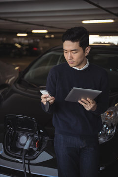 Man using digital tablet and mobile phone while charging electric car