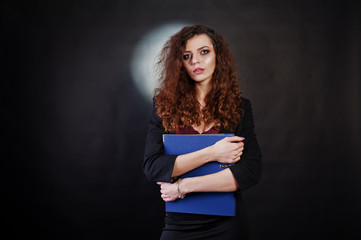 Brunette curly haired girl in black office jacket with skirt, on bra with folder of documentts at hands on studio against black background. Sexy businesswoman.