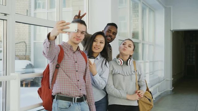 Group of four multi-ethnic positive male and female students are standing in wide corridor. Hipster gut is holding smartphone making selfie of them all