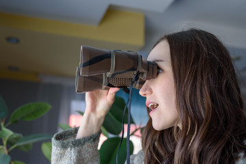 Young woman spying out the window looking through binoculars