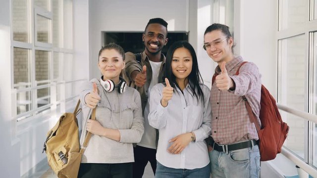 Portrait of four positive multi-ethnic male and female students standing in spacious white corridor in university looking at camera showing thumbs up