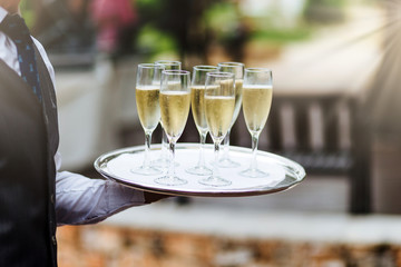 Unrecognizable waiter in formal clothing holds tray with glasses of champagne, carries drinks on festive table to guests, supplies good service for visitors. Hospitality, service and drinking concept