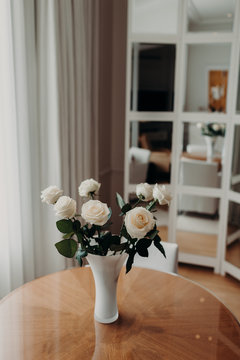 Vertical shot of beautiful bouquet of white roses in vace stands on wooden round table against cozy home interior. Wonderful roses with pleasant odour