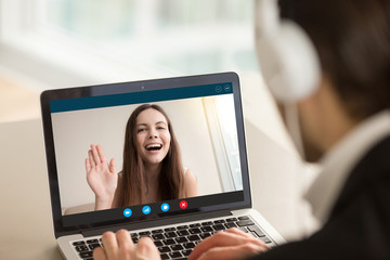 Fototapeta na wymiar Young girl waving from laptop screen at man in headphones. Happy teen greeting her boyfriend, friends exchange and learn foreign languages, couple communicating via video call app. View over shoulder.