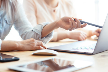 Close up shot of two female businesswomen working with hands on a laptop keyboard in an office in sunlight at a wooden table and one of them showing a pen on the display